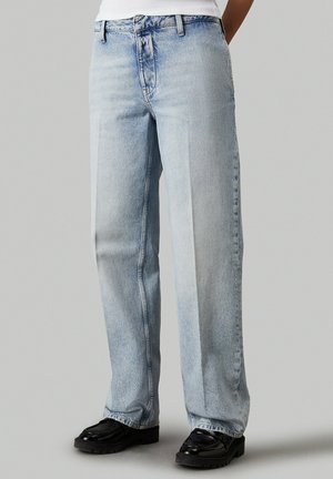 Persoon draagt lichtblauwe rechte jeans en zwarte loafers, staand tegen een effen grijze achtergrond.