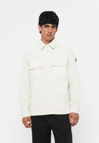Camicia in corduroy bianco con chiusura a bottone, due tasche sul petto e una finitura testurizzata. Presenta un colletto e un piccolo logo sul maniche.
