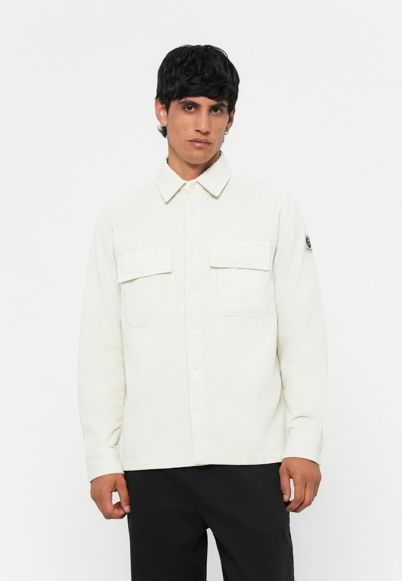 Camicia in corduroy bianco con chiusura a bottone, due tasche sul petto e una finitura testurizzata. Presenta un colletto e un piccolo logo sul maniche.