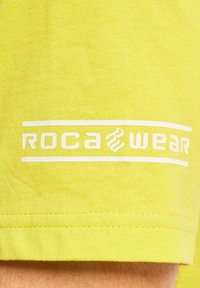Neon geltonos spalvos medvilninė marškinė su lygiu paviršiumi, ant kurių uždėtas baltas spaudinys "ROCA WEAR" ir horizontalaus dizaino elementas.
