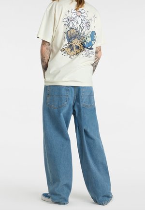 T-shirt jaune clair avec un graphique de crâne floral au dos, associé à un jean en denim bleu à coupe décontractée et des baskets bleues.