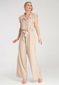 Beżowy kombinezon bez rękawów z zapinanym przodem, dwiema kieszeniami na piersi i paskiem w talii. Miękka tkanina; szerokie nogawki i zwiewna faktura.