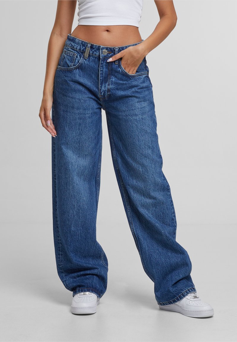 Lange denim jeans in een middelblauwe wassing, met een hoge taille, vrouwelijke zakken en subtiele vervaging. Gecombineerd met witte sneakers.