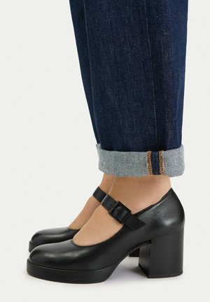 Czarne skórzane buty Mary Jane na szerokich obcasach, noszone z podwiniętymi ciemnoniebieskimi dżinsami.