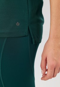 Grünes Sportoberteil aus strukturiertem Stoff, mit seitlichem Schlitz und Logodetail, kombiniert mit passenden grünen Leggings. Nahaufnahme einer Hand auf dem Stoff.