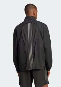 Black windbreaker jacket featuring a textured back with a rounded cut and three reflective stripes down the center. The design includes a high collar.