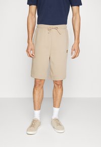 Shorts en sweat beige avec taille élastique et cordon de serrage, dotés de poches latérales et d'un petit logo sur la cuisse gauche. Portés avec une chemise navy.