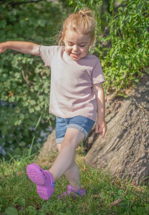 Kind, der ein hellrosa geripptes Kurzarmshirt, Jeansshorts und lila Slipper trägt, steht auf Gras neben einem Baum.