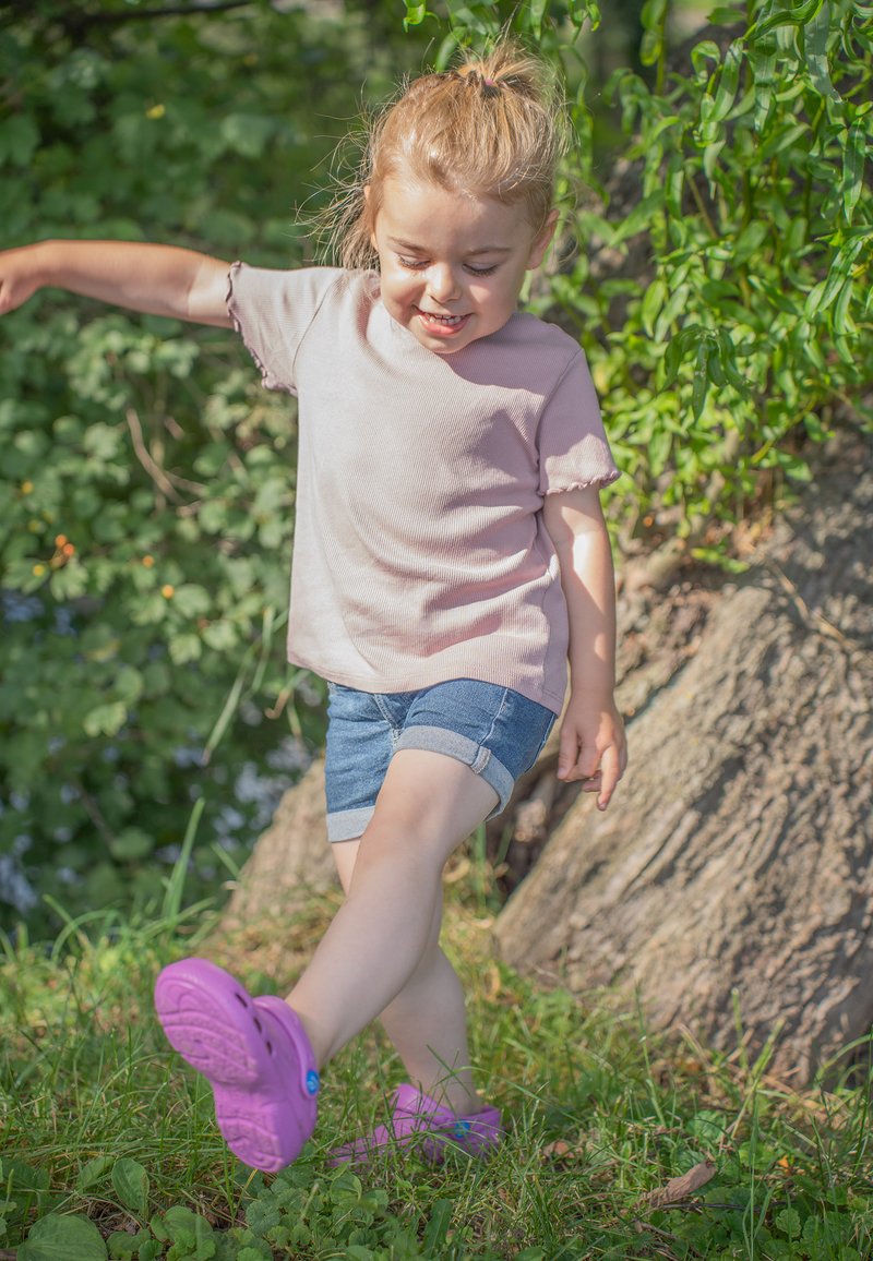 Child wearing a light pink ribbed short-sleeve shirt, denim shorts, and purple slip-on shoes, standing on grass near a tree.