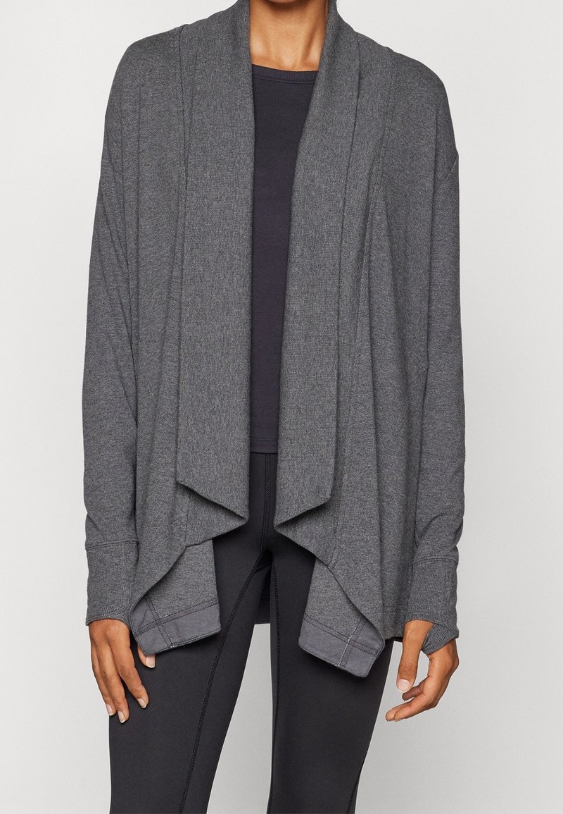 Personne portant un cardigan gris foncé ample et ouvert sur le devant, par-dessus un haut noir et des leggings noirs, debout devant un fond clair uni.
