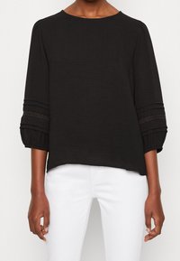 Blouse noire texturée à manches trois-quarts avec des accents volantés et des détails en dentelle, assortie à un bas blanc.
