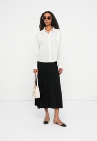 Biała koszula zapinana na guziki z subtelnym kwiatowym wzorem, czarna spódnica plisowana, beżowa torebka i cieliste baleriny z czarnym czubkiem, stylizowane z okularami przeciwsłonecznymi.