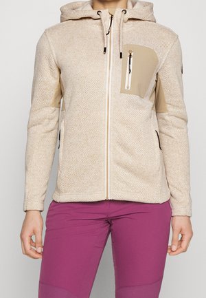 Beiger, texturierter Hoodie mit Reißverschluss, vorderer Brusttasche und Kordeln, getragen mit leuchtend violetten, enganliegenden Hosen an einer stehenden Person.