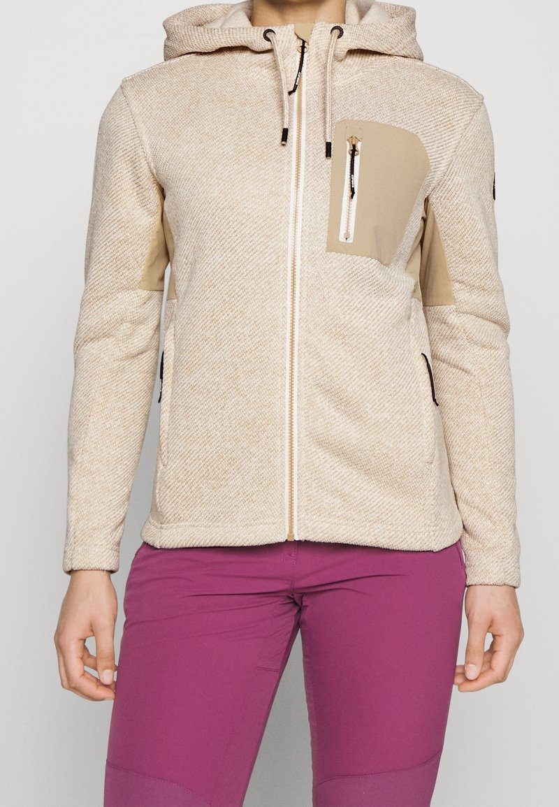 Sweat à capuche beige zippé texturé avec poche poitrine avant et cordons, porté avec un pantalon ajusté violet vif sur une personne debout.