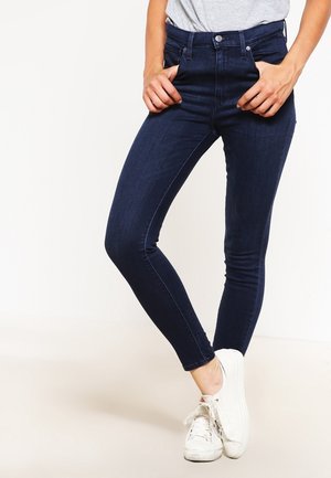 Jeans de tiro alto, corte ajustado en denim azul oscuro con una textura suave. Cierre de botón y cremallera, combinados con zapatillas blancas.