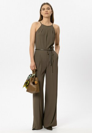 Woman wearing olive green sleeveless jumpsuit with belt holding brown handbag with patterned scarf, standing against white background.