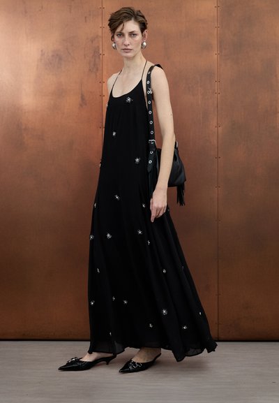 Black long dress with silver embellishments, spaghetti straps, and flowy fabric; paired with black handbag and pointed mules.