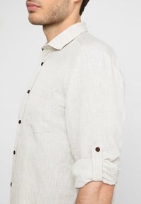 Camisa de lino de color claro con cuello abotonado, mangas remangadas, un bolsillo en el pecho y botones marrón oscuro. Textura suave, ajuste relajado.