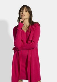 Vestido suéter de cuello en V en rosa brillante, hecho de una tela suave. Cuenta con mangas largas con puños acanalados y un corte relajado, con un detalle de tejido drapeado.