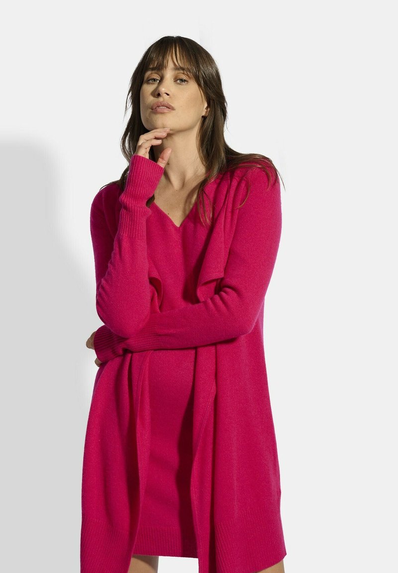 Vestido suéter de cuello en V en rosa brillante, hecho de una tela suave. Cuenta con mangas largas con puños acanalados y un corte relajado, con un detalle de tejido drapeado.