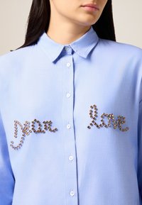 Camisa de botones azul claro con collar que presenta gemas decorativas marrones formando las palabras "Querido" y "Amor" en el frente. Textura suave.