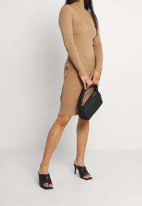 Robe col roulé côtelée couleur sable avec détail de boutons sur le côté, associée à un sac à main tressé noir et des mules noires à bout ouvert, posant devant un fond blanc.