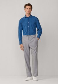 Uomo che indossa una camicia button-up in denim blu, pantaloni grigi chiari su misura con coulisse e sneaker bianche, in piedi contro uno sfondo neutro.