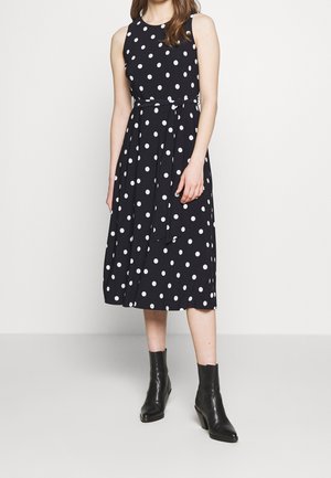 Femme portant une robe midi noire sans manches à pois blancs avec une ceinture nouée à la taille, associée à des bottines noires.
