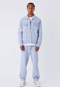 Chaqueta de mezclilla azul claro y pantalones a juego con un patrón texturizado. Camiseta blanca debajo. Modelo posando contra un fondo blanco.