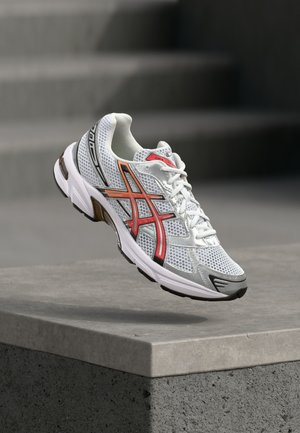 Zapatilla deportiva blanca con parte superior de malla, acentos rojos y negros, suela de goma gris y puntera reforzada. Ligera, diseñada para ofrecer soporte y transpirabilidad.