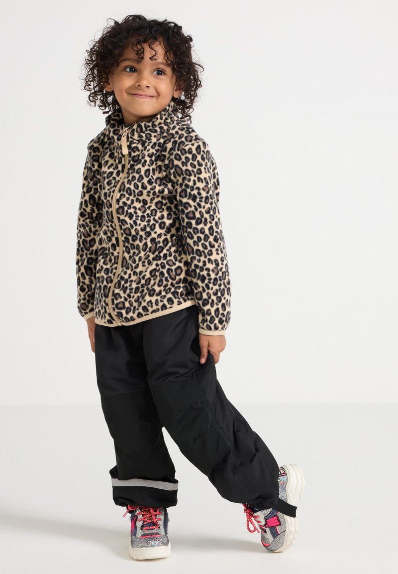 Enfant aux cheveux bouclés portant une veste zippée à motif léopard, un pantalon noir et des baskets colorées, debout avec un pied légèrement levé.