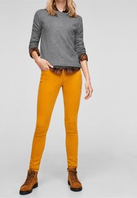 Frau in senfgelben engen Hosen, karamellfarbenen Schnürboots, einem grauen Pullover mit hochgerollten Ärmeln und einem karierten Hemd darunter.