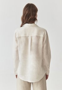 Světle béžová lněná košile s límečkem, dlouhými rukávy a zaobleným lemem. Obsahuje zádový yoke a detail na rukávech pro ohrnutí s knoflíkem.