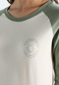 Camicia a maniche lunghe con il corpo di un beige chiaro e maniche raglan verdi. Presenta una grafica circolare di uno squalo in verde tenue nella zona del petto.