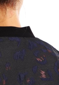 Svart texturerad tyg med lila och metalliska ros mönster; ribbad svart krage; bakvy som visar detaljerad design och sömnad.