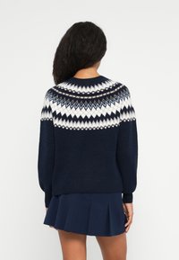Frau mit langen dunklen Haaren, die einen marineblauen Pullover mit weißen, beigen und hellblauen geometrischen Mustern sowie einen marineblauen plissierten Rock trägt.