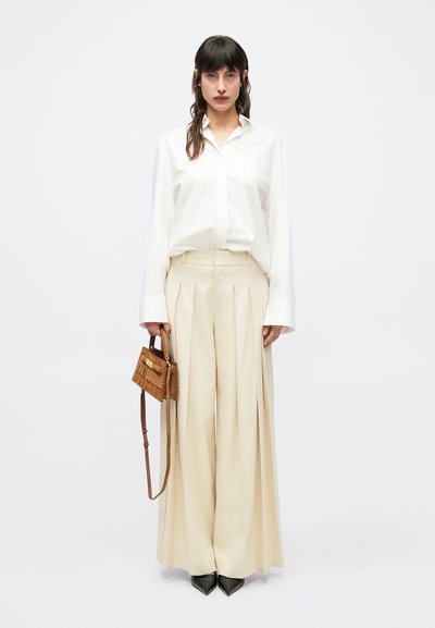 Valge nööpidega särk, laiad beežid volditud püksid, mustad terava ninaga kingad ja väike pruun tutikujuline õlakott.