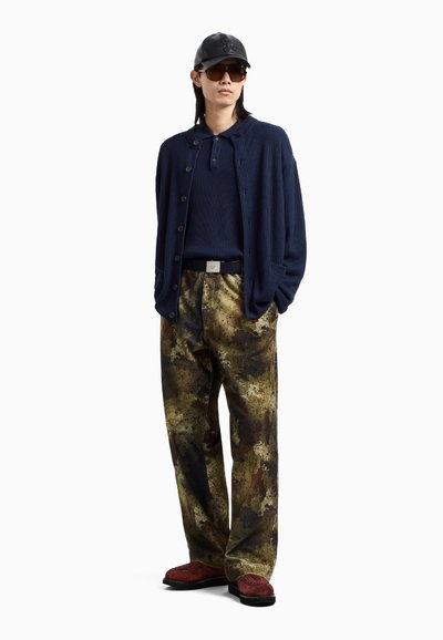 Cardigan a coste blu navy sopra un polo coordinato, abbinato a pantaloni a motivi oliva e marroni. Gli accessori includono un cappellino nero e occhiali da sole.