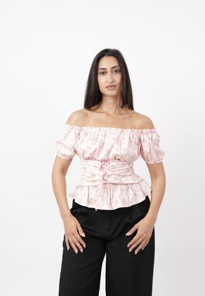 Mujer con cabello largo y oscuro que lleva una blusa rosa con flores y escote descubierto en los hombros, y pantalones negros, de pie frente a un fondo blanco liso.