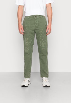 Man wearing green distressed straight-leg pants, white sneakers, and a white t-shirt, standing against a plain white background.