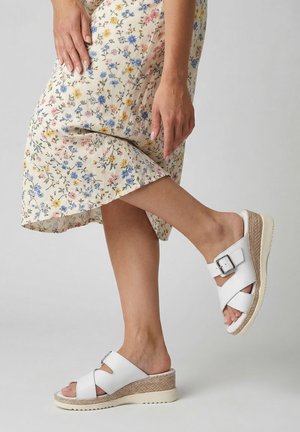 Frau trägt weiße Keilsandaletten mit Schnallen und einen geblümten Midirock vor einfarbigem Hintergrund.
