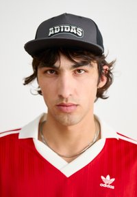 Μαύρο καπέλο με το λογότυπο "ADIDAS", κόκκινο πουκάμισο με κάθετες ρίγες και λευκό γιακά, ασημένιο κολιέ, ουδέτερο φόντο.