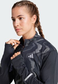 Black athletic jacket featuring a half-zip collar, patterned with white dots and textured sleeves. Long sleeves have thumbholes and reflective accents.