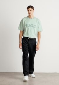 Moški nosi oversize majico mint zelenkaste barve z napisom "HUGO", črne hlače z pasom in bele superge, medtem ko hodi po notranjosti na sivi talni oblogi.