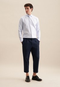 Camicia azzurra chiaro con maniche lunghe, abbinata a pantaloni cropped blu navy e scarpe nere con lacci, in piedi di fronte a uno sfondo neutro.
