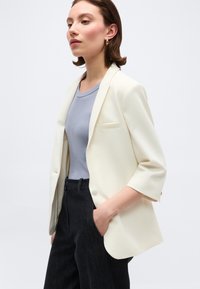 Γυναίκα που φοράει εκρού σακάκι με μανίκια ως τον αγκώνα, ανοιχτό μπλε μπλουζάκι, μαύρο παντελόνι και χρυσούς κρίκους σκουλαρίκια, στέκεται με το ένα χέρι στην τσέπη.