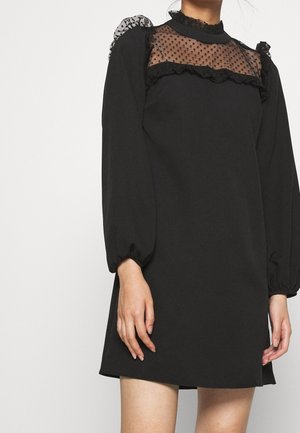 Černé šaty s průsvitným živůtkem z puntíkované síťoviny s volánky, dlouhými nafouknutými rukávy a jednoduchým áčkovým střihem.