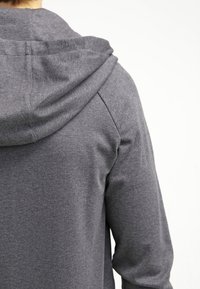 Sweat à capuche gris en tissu texturé, doté d'une capuche à cordon et de manches raglan. Vue rapprochée de dos, mettant en valeur les détails de couture.