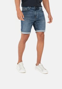 Denim short met een medium blauwe kleur, opgerolde zomen, vijf zakken en contrasterende gele stiksels, gecombineerd met witte sneakers.