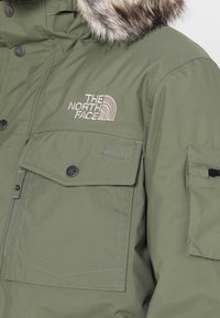 Veste d'hiver vert olive avec capuche bordée de fourrure, poche avant à rabat, et logo "The North Face" brodé sur la poitrine.